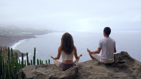 At-the-peak-of-a-mountain,-a-man-and-woman-sit-on-stones,-meditating-with-raised-hands,-overlooking-the-ocean,-and-practicing-serene-breaths